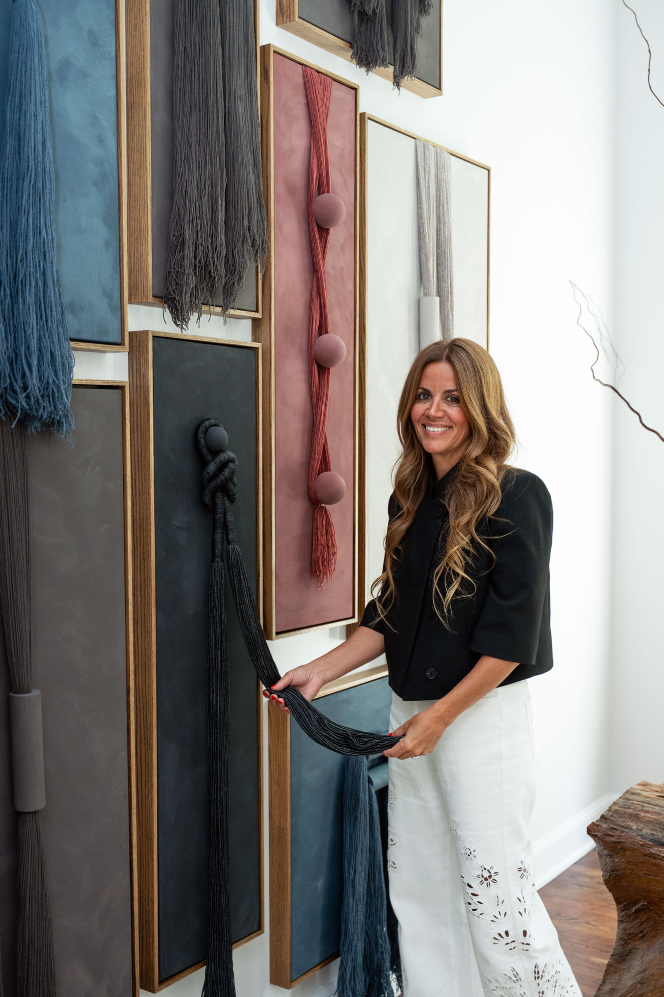 Artist Patchi Cancado standing beside her collection of original contemporary canvas and yarn artworks, featuring sculptural fiber details, dimensional textures, muted colors, and handcrafted wood frames.