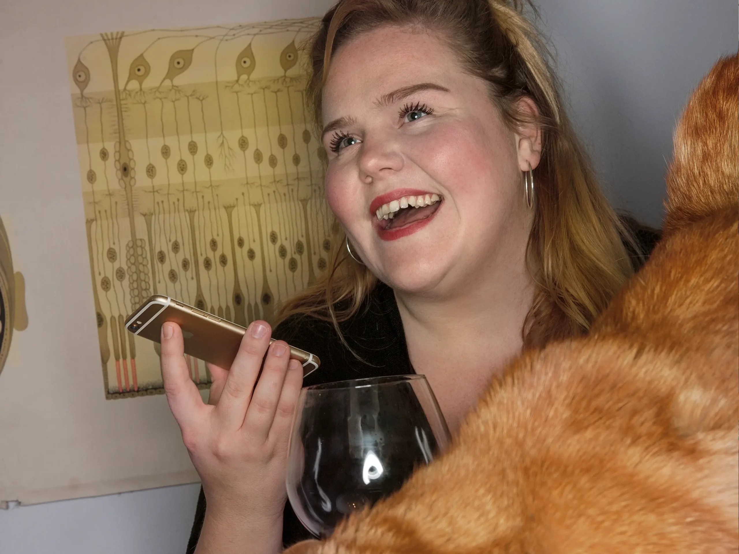 Porträtt av Linnea Wikblad i ett kök, hon har en mobiltelefon i handen, skrattar, framför henne spår av katt och vinglas.