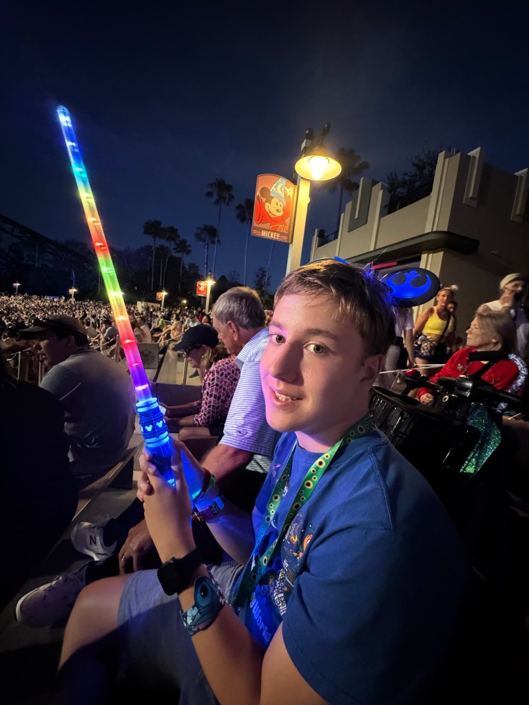 A young boy sitting in a crowded area at Disney World, smiling and holding a brightly colored light-up toy. He is wearing a blue shirt and has a green identification wristband.
