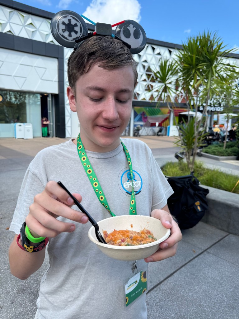 A young boy wearing themed ears and a lanyard is enjoying a bowl of food at Disney World, smiling as he prepares to take a bite.
