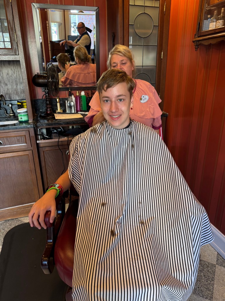 A young boy sitting in a barber chair wearing a striped cape, smiling while a hairstylist prepares for a haircut. Reflections of other customers and staff are visible in the mirror.