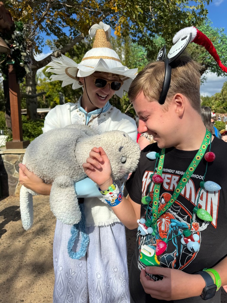 A young boy smiles while holding a fluffy gray toy and interacting with a friendly staff member wearing a hat and sunglasses in a theme park setting.