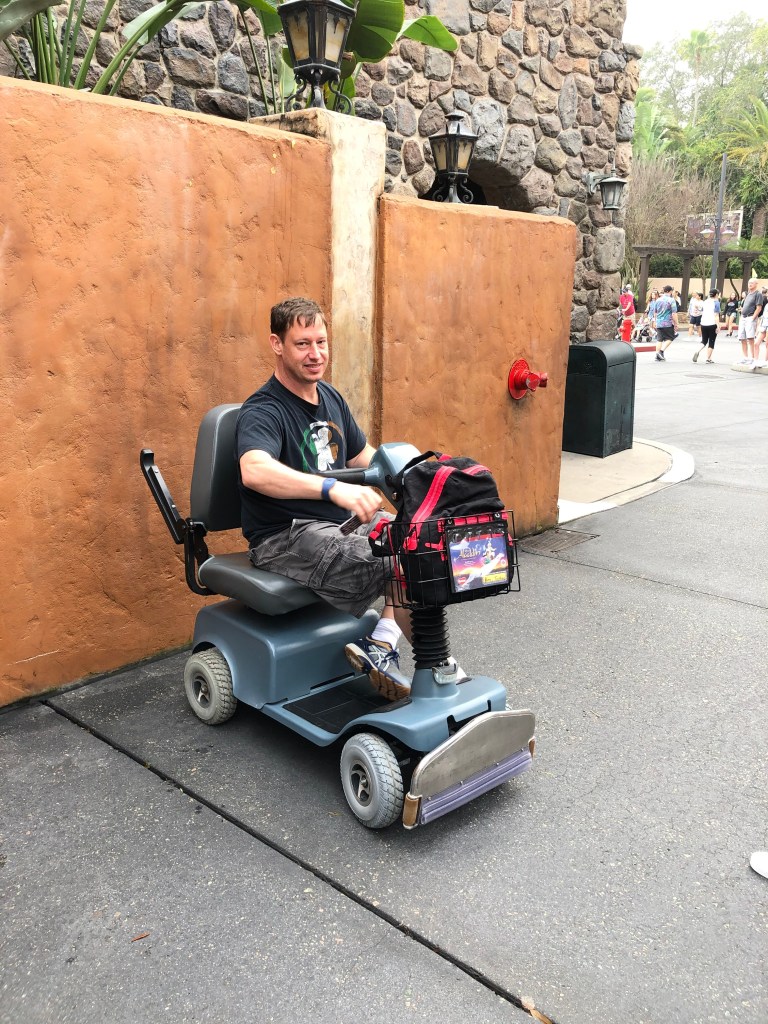 A man sitting on a mobility scooter in a Disney theme park, smiling and enjoying his surroundings.