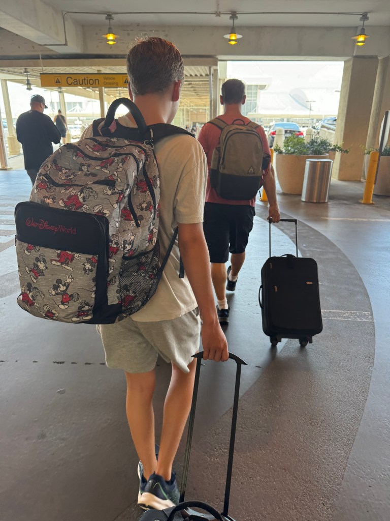 A young person walks through an airport with a Disney-themed backpack, pulling a suitcase, while another traveler follows behind.
