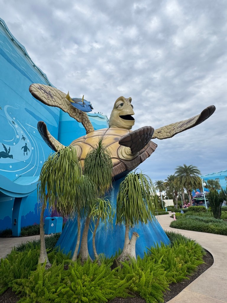 A large, colorful statue of a turtle with a friendly expression, situated in front of a blue wall adorned with aquatic-themed designs. Lush green plants surround the statue, adding to the vibrant atmosphere.