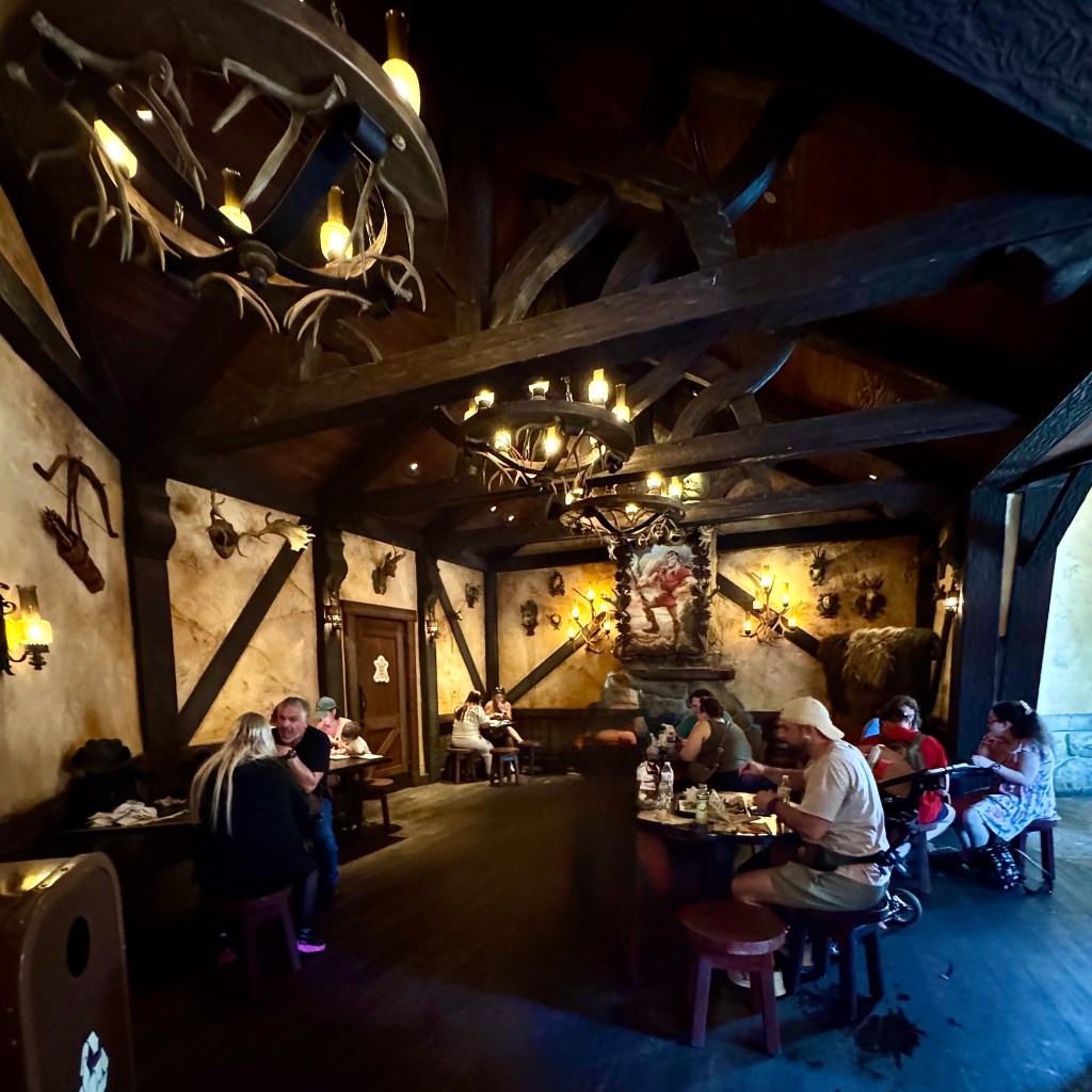 Interior of Gaston's Tavern at Magic Kingdom, featuring rustic decor, wooden beams, and guests dining at tables.