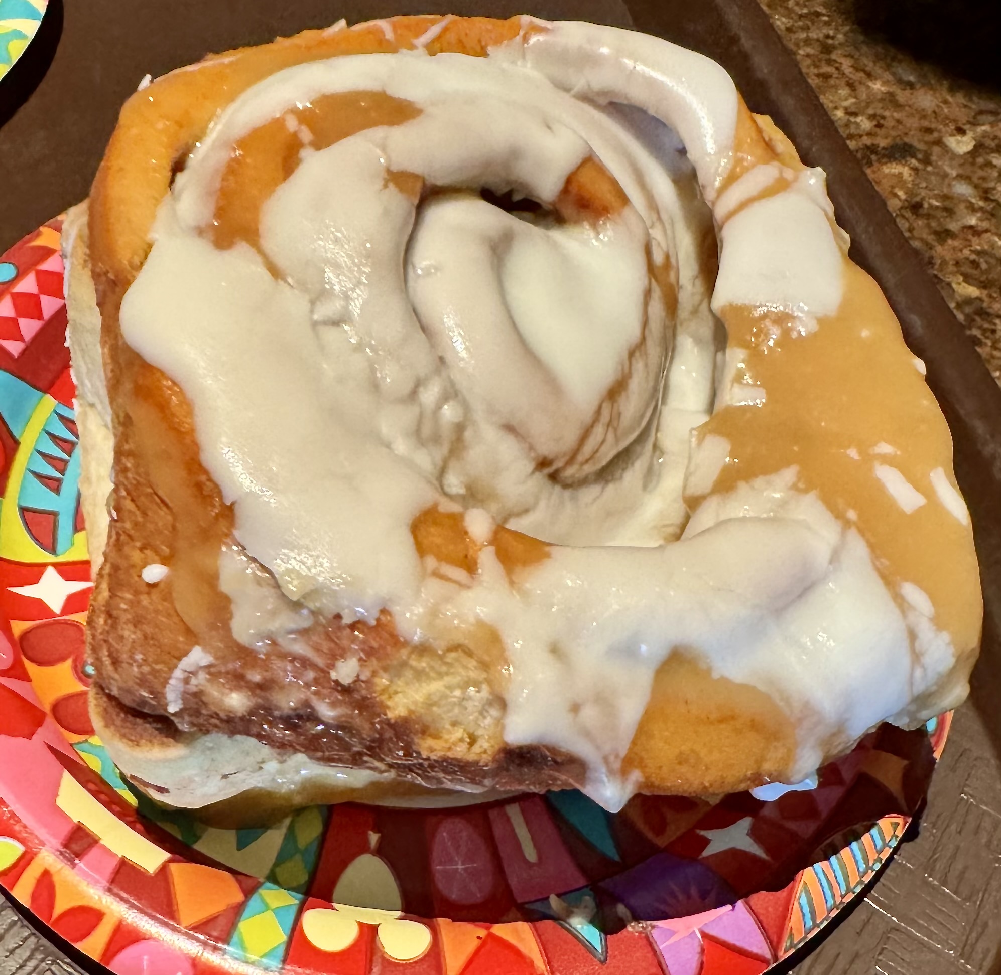 A large cinnamon roll topped with white icing, displayed on a colorful patterned paper plate.
