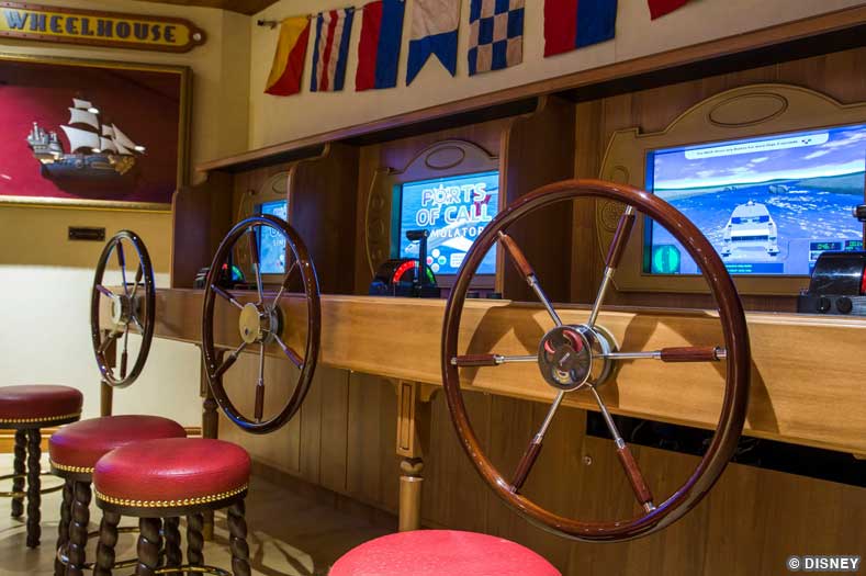 Cruise ship simulators with red stools, large ships wheels, and video screens with docking controls, in the Wheelhouse of the Oceaneer Lab on Disney Magic