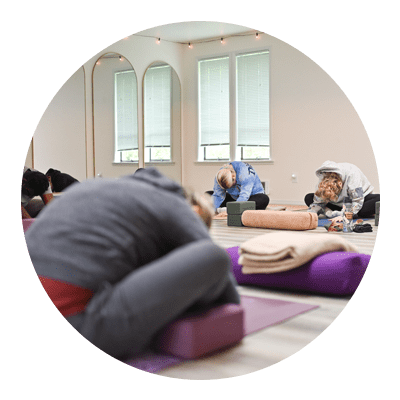 A small group yoga class at Mahad practicing a seated forward fold stretch in a bright studio with wooden floors. Students sit on mats using bolsters and blocks for support, with large arched mirrors and string lights along the wall creating a calm atmosphere.