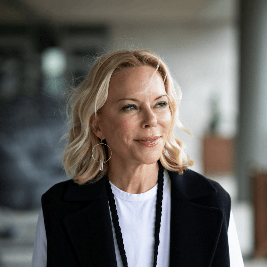Portrait of a smiling blonde woman in a white shirt and black vest, looking to the right in a bright indoor corridor.