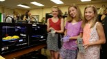 Three middle school girls hold e-NABLE hands next to a makerbot printing a hand