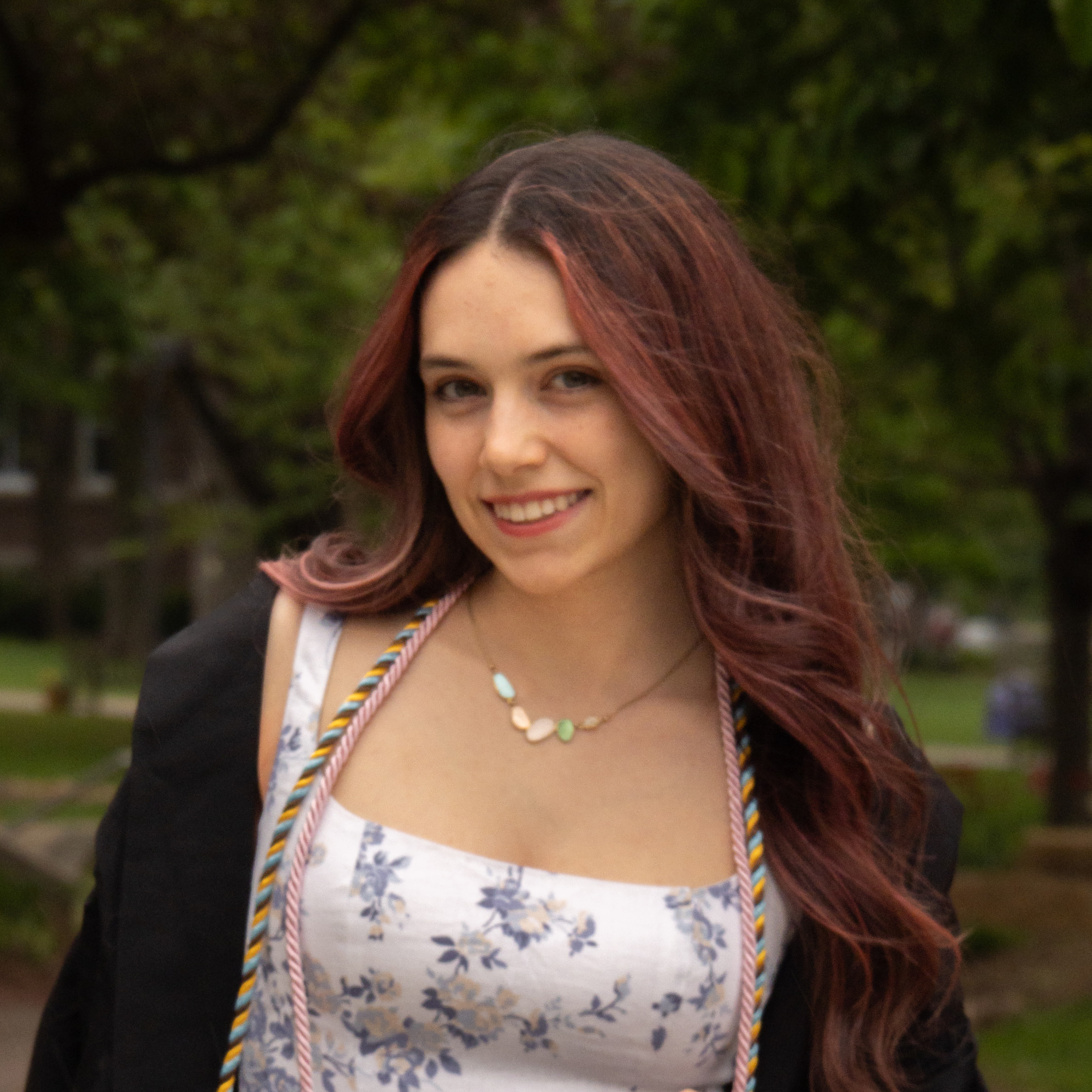 Pictured is a white woman with red, curled hair wearing a white dress with blue flowers, two graduation cords, and a black graduation gown. She has dark green trees behind her.