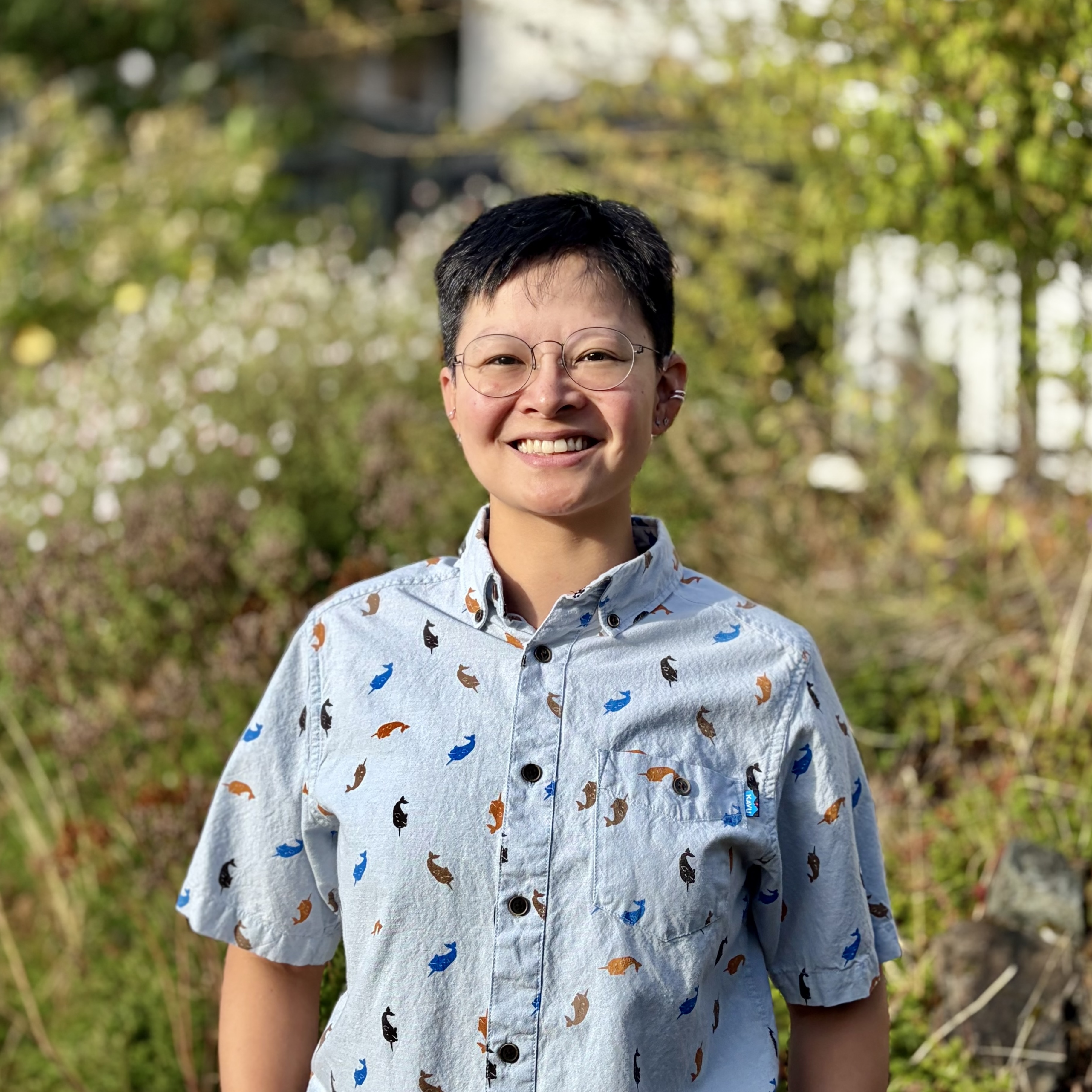 Portrait of Sev Huffman, a smiling Asian person with short black hair and round glasses, standing outdoors in a sunlit garden and wearing a light blue button-up shirt with a colorful print.