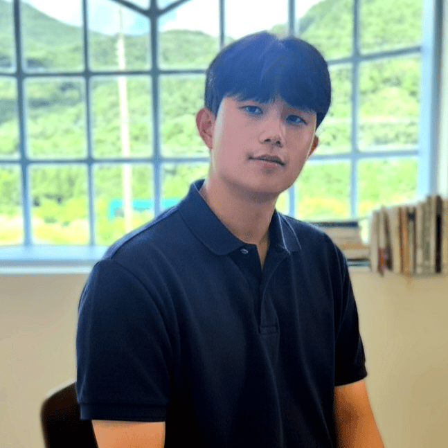 Gyeongdeok Kim, a man with short black hair and wearing a navy polo shirt, sits facing the camera in a bright room with large windows overlooking green hills and books beside him.