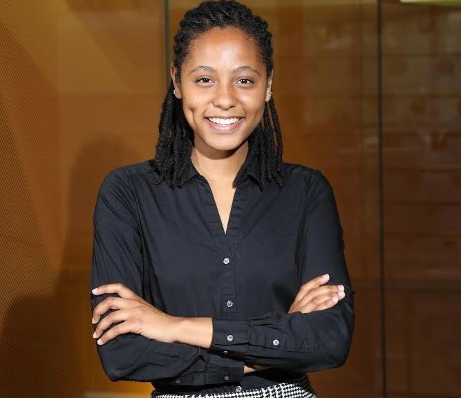 Aaleyah, a Black woman with black locs pulled back, wearing a black button up shirt, smiling at the camera.