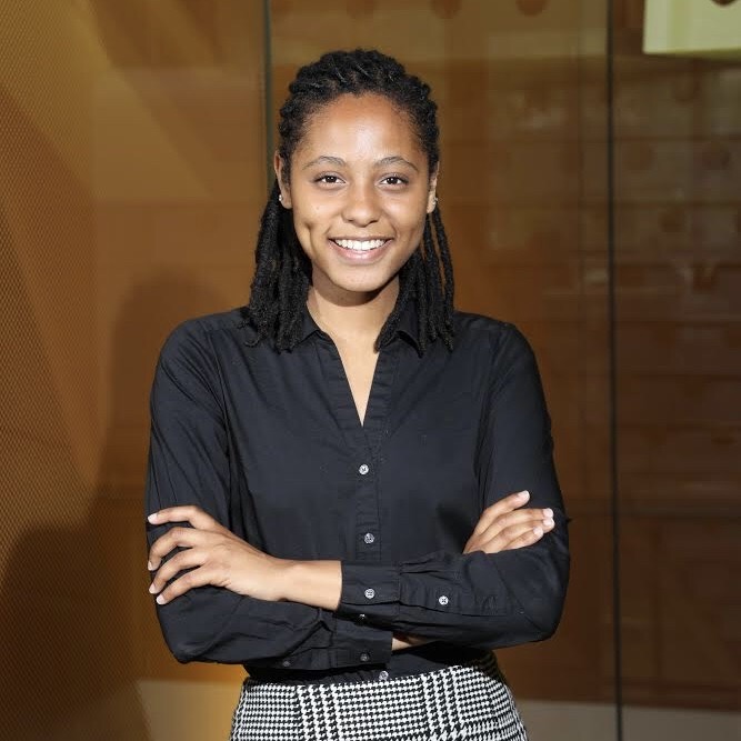 Aaleyah, a Black woman with black locs pulled back, wearing a black button up shirt, smiling at the camera.