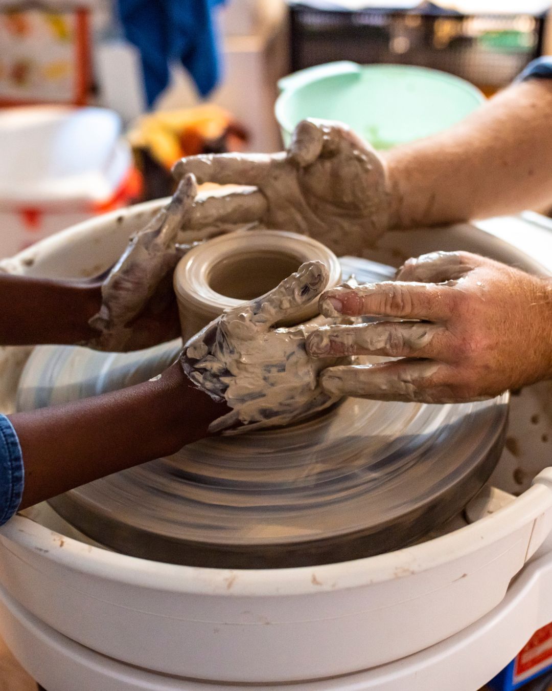 Two people side by side at pottery wheels, shaping clay with their hands, smiling and focused. The words “Stronger Together” appear over the image, highlighting the spirit of creative collaboration.
