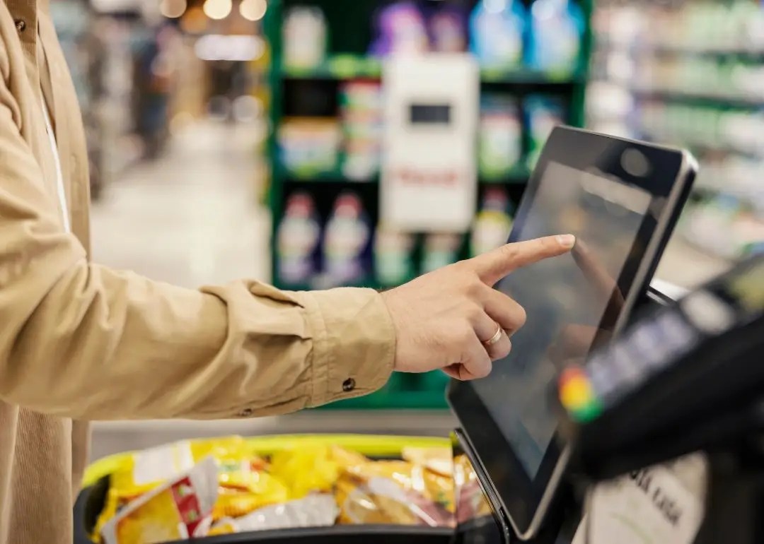 Person uses a touchscreen checkout tablet at a grocery store self-checkout, with snacks in the foreground.