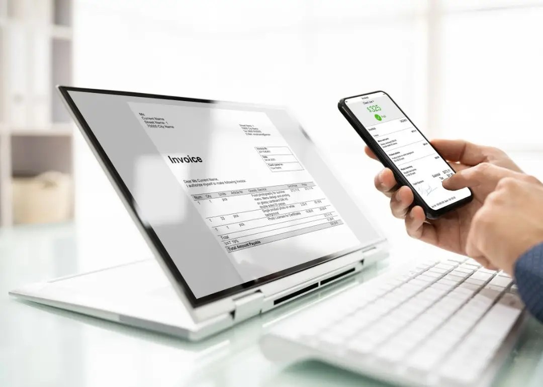 Invoice displayed on a laptop screen while a hand holds a smartphone showing a financial app beside it.