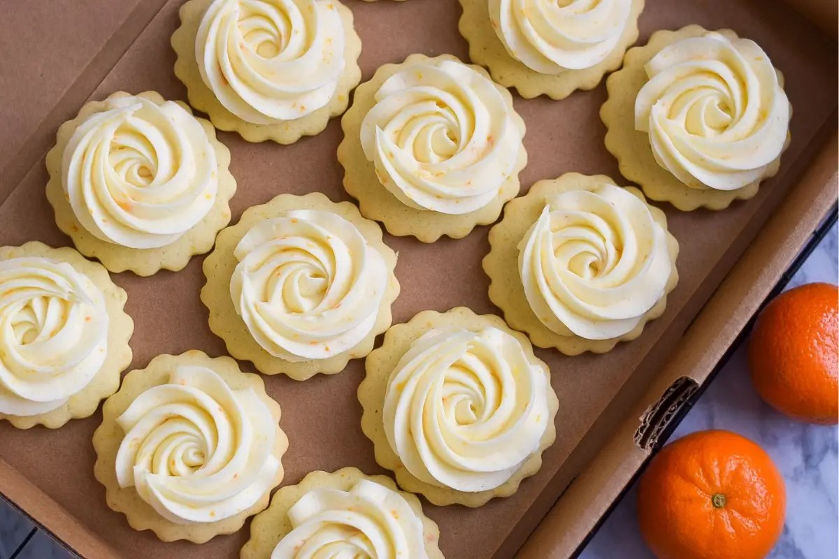 Tray of lemon-tart cookies with ivory buttercream swirls in a cardboard box, with two oranges visible at the side.