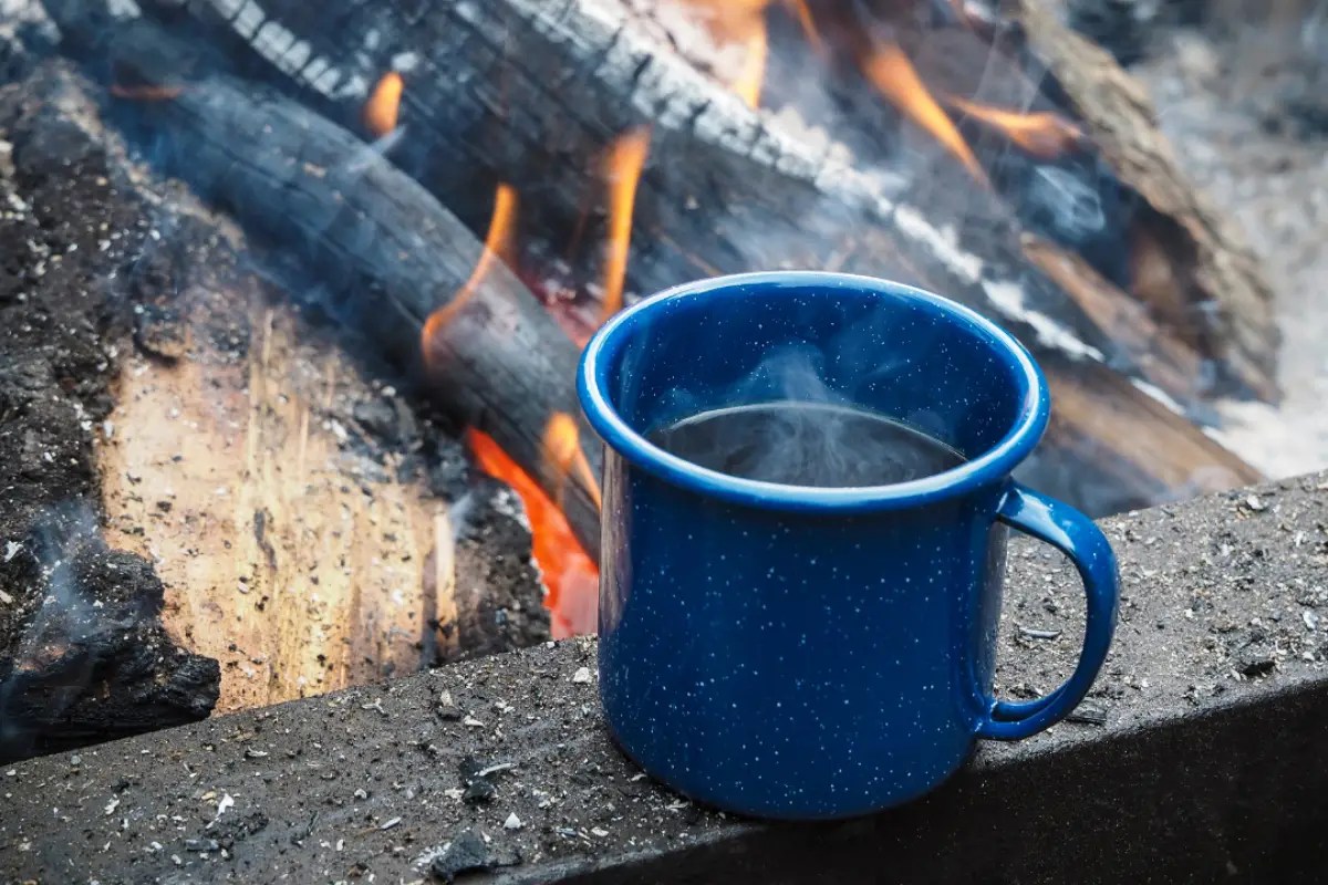 Blue enamel mug with steam by a campfire on a rocky surface; glowing logs in the background.