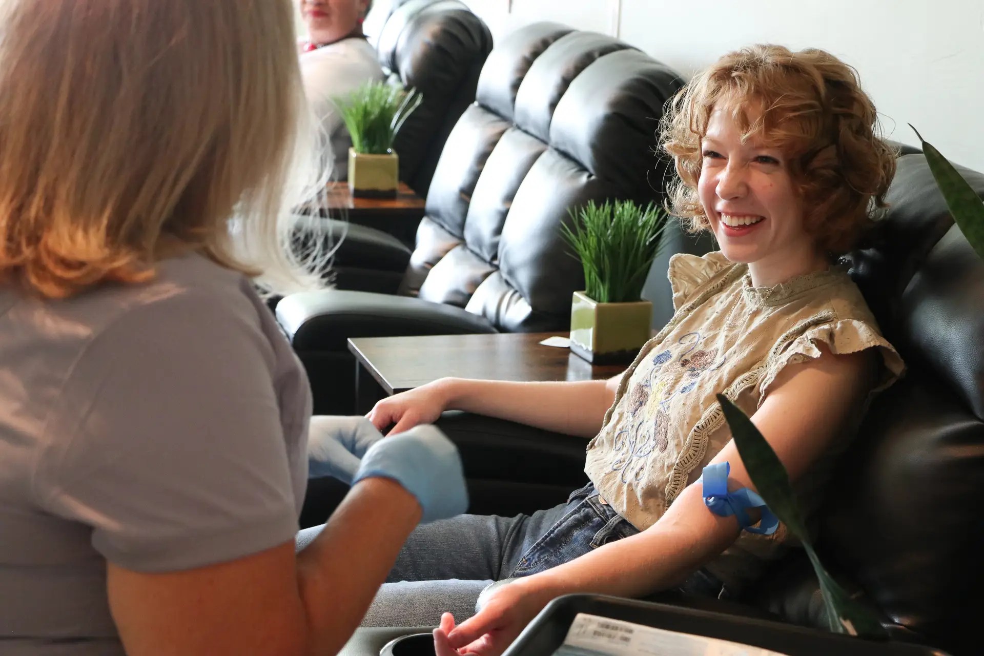 Woman smiles while donating blood in a clinic, seated in a reclining chair with a nurse/staff preparing her arm.
