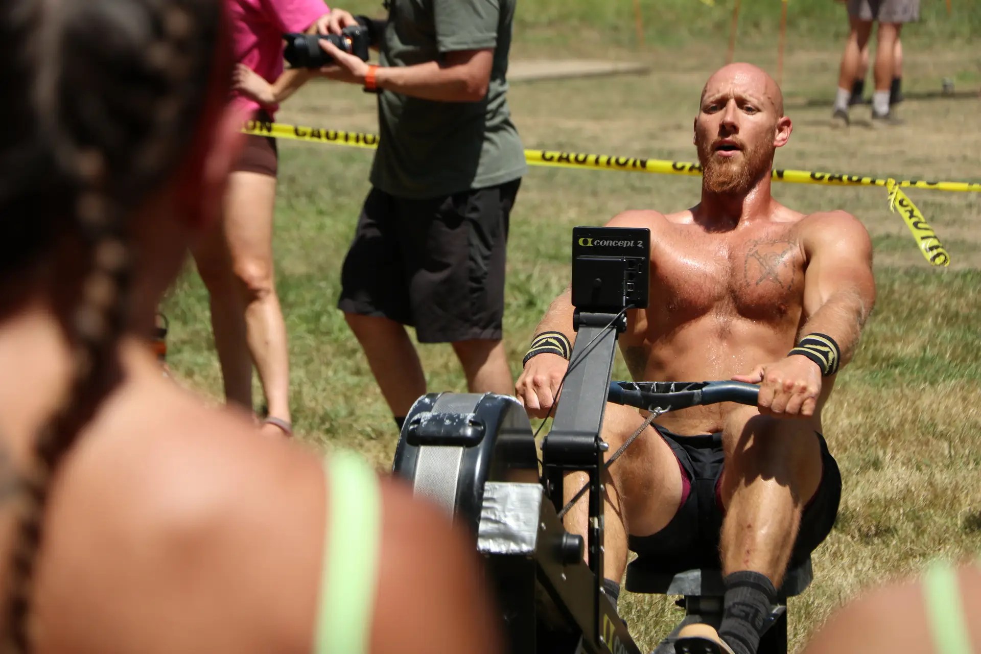 Shirtless bearded man riding a stationary bike outdoors at a sunny event, with spectators and yellow caution tape behind him.