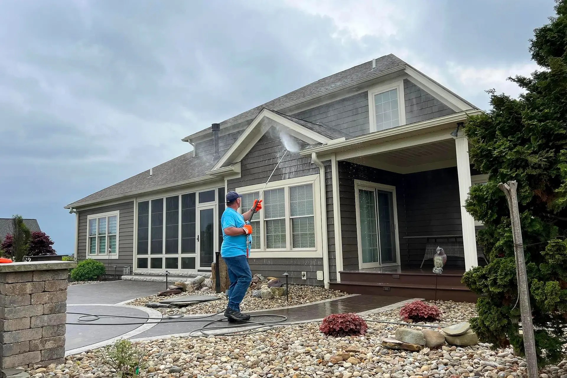A man in a blue shirt and cap pressure-washes the front of a gray house with a long wand, spraying water near the siding.