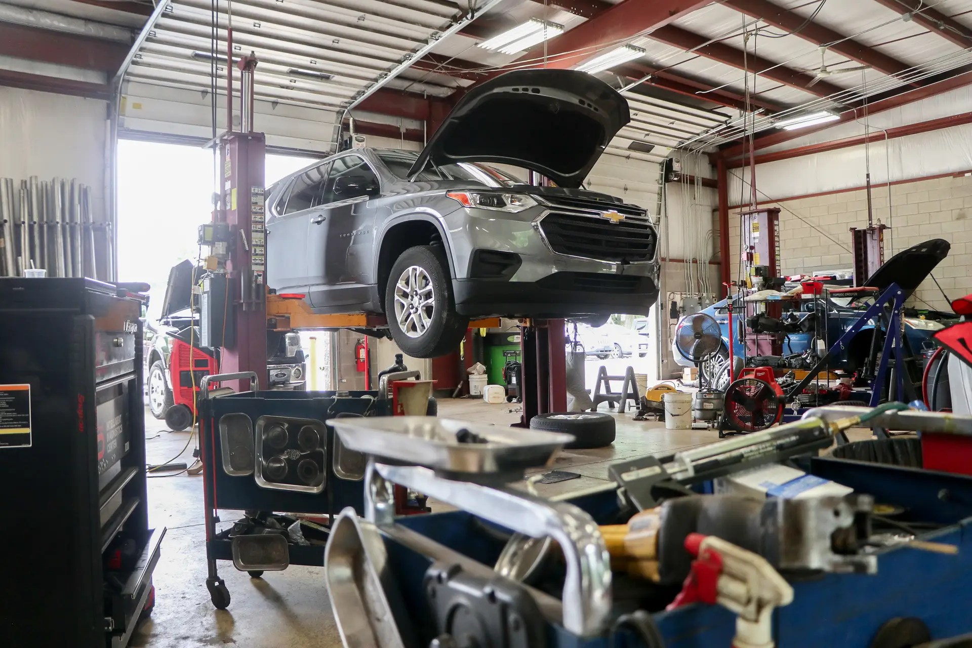 Silver Chevrolet SUV on a hydraulic lift with its hood open inside an auto shop, tools and equipment scattered around