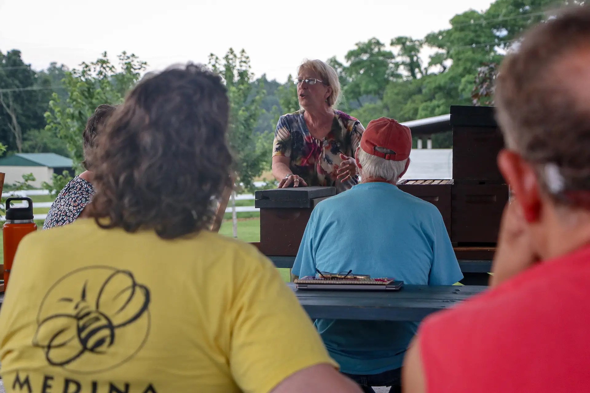 Older woman speaks at an outdoor gathering behind a podium; audience sits, foreground shows a person in a yellow bee-graphic shirt.