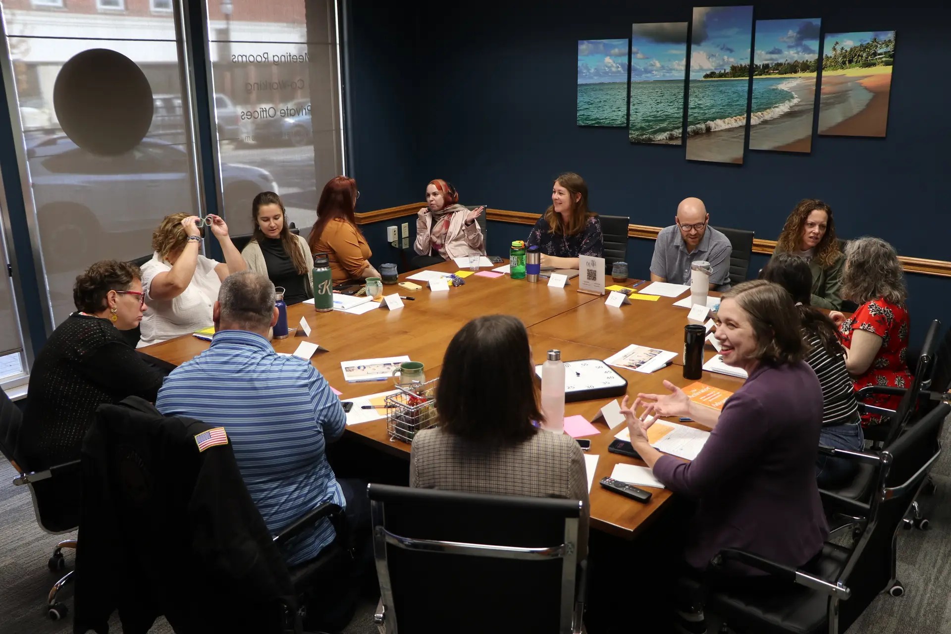 Group of adults around a large conference table in a meeting room, chatting and taking notes with papers and name cards on the table.