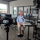 Man in a light blue shirt sits on a stool in a television studio, surrounded by cameras, lights, and monitors.