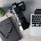 DSLR camera with a large lens rests on a notebook beside a laptop, glasses, and a small plant on a wooden desk.