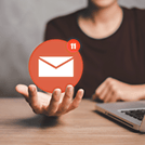 Person holds an orange envelope notification icon above an open hand, signaling a new email message (11 new) and a nearby laptop on a desk.