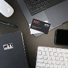 Desk workspace with a laptop, white keyboard, white mouse, notebook, and a small card on a dark desk surface.