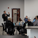 Facilitator standing with a microphone at the front while adults sit around a conference table in a workshop room.