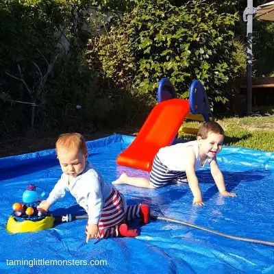 Two toddlers playing on a DIY splash pad in the backyard, crawling on a blue tarp with water spraying nearby and a small slide in the background.