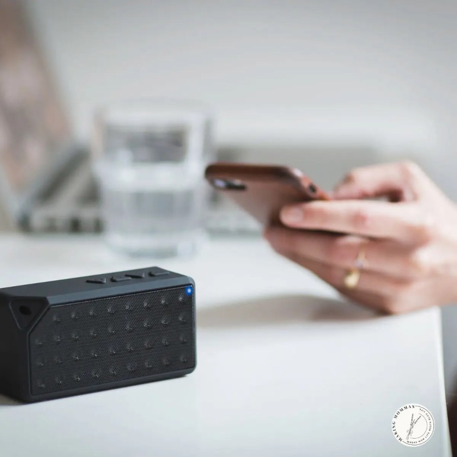 Small black Bluetooth speaker on a white desk with a blurred background of a person using a smartphone and a laptop, representing a modern and functional home office setup.