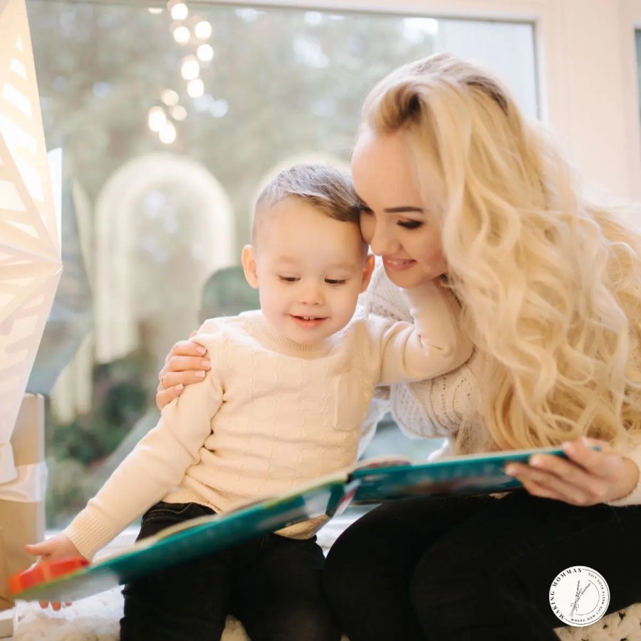 A mother and her young child sitting by a window, smiling as they read a colorful book together in cozy winter sweaters.