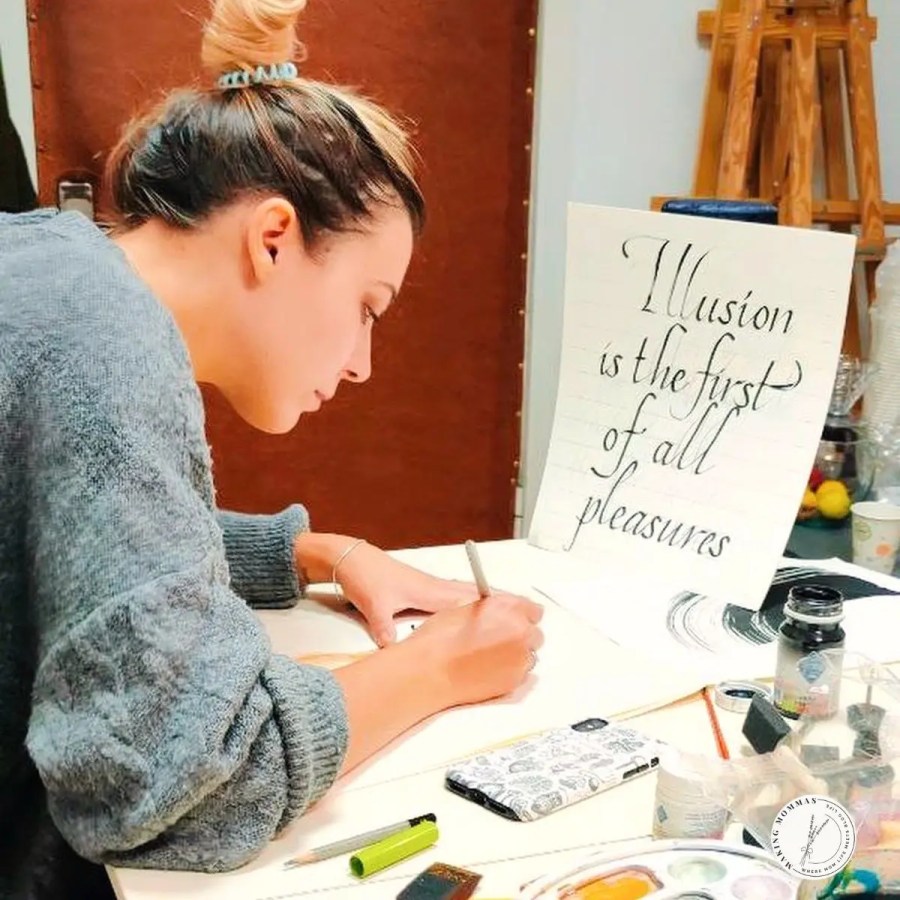 A woman practicing calligraphy at a desk with brushes, ink, and pens, writing a quote on a sheet of paper that reads “Illusion is the first of all pleasures.”
