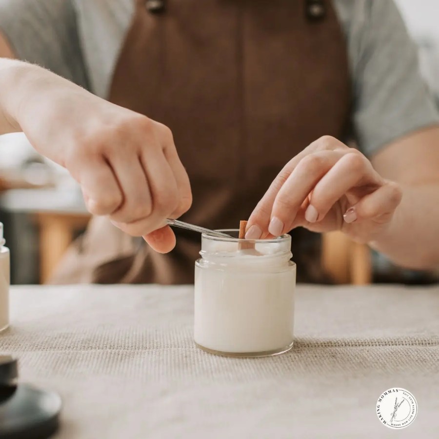 A close-up of a person making candles by hand, carefully adjusting a wooden wick inside a glass jar filled with creamy white wax on a beige table.