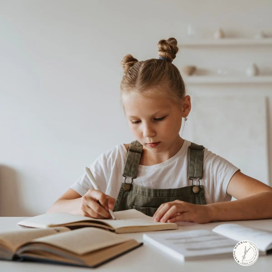 Young girl with braided buns wearing overalls, sitting at a desk and writing in a notebook while doing homework.