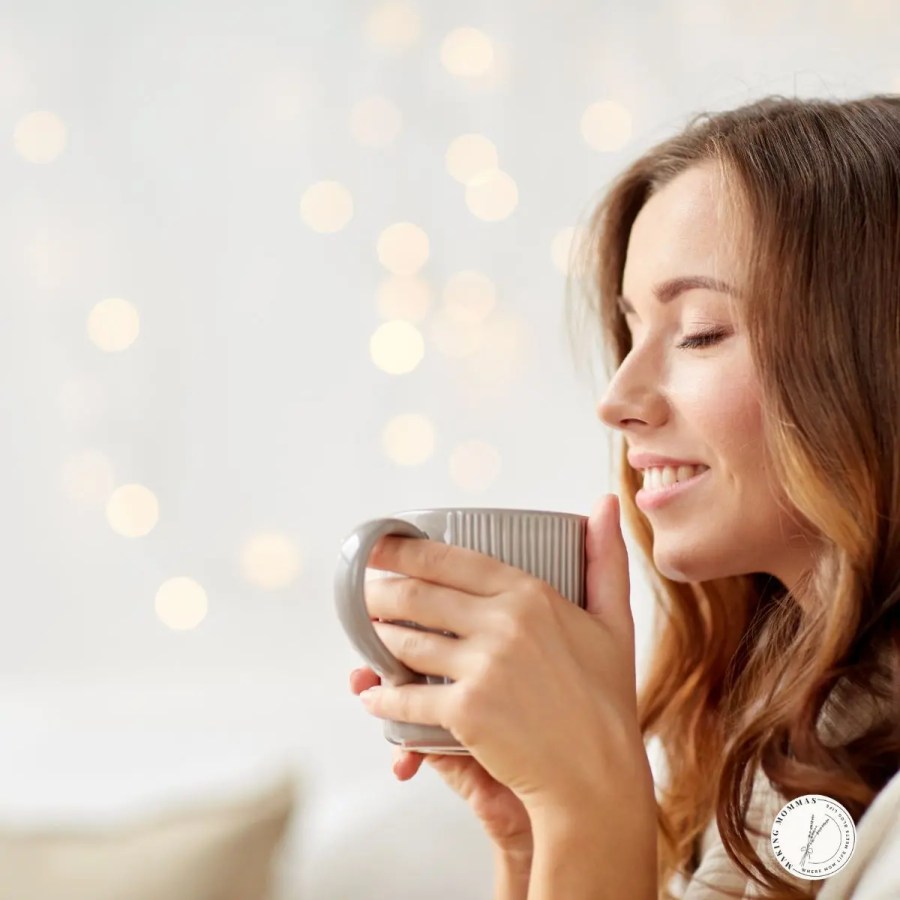 Smiling woman holding a warm mug close to her face with eyes closed, enjoying the aroma of freshly brewed coffee in a cozy, softly lit home setting.