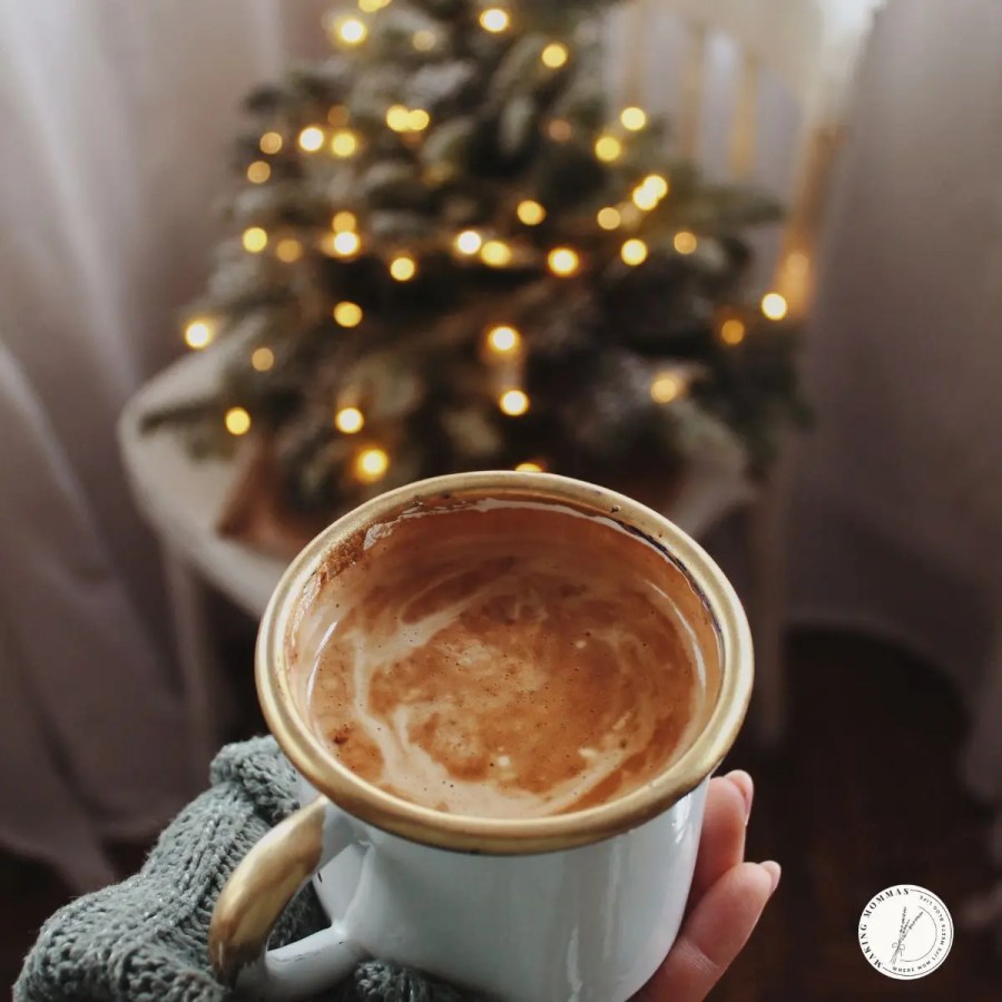 Hand holding a cozy mug of coffee with frothy swirls, in front of a softly lit Christmas tree, capturing a warm and comforting holiday moment for work-from-home moms.