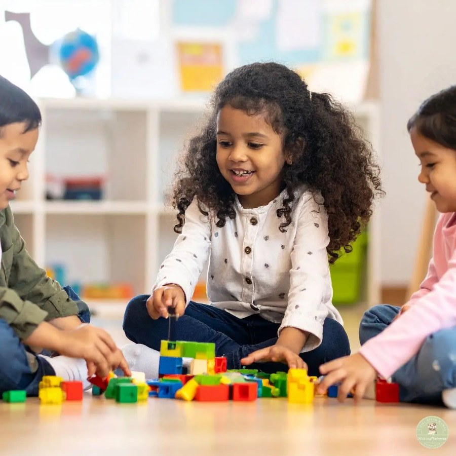 Three young children sitting together on the floor, smiling and building with colorful blocks during collaborative play.