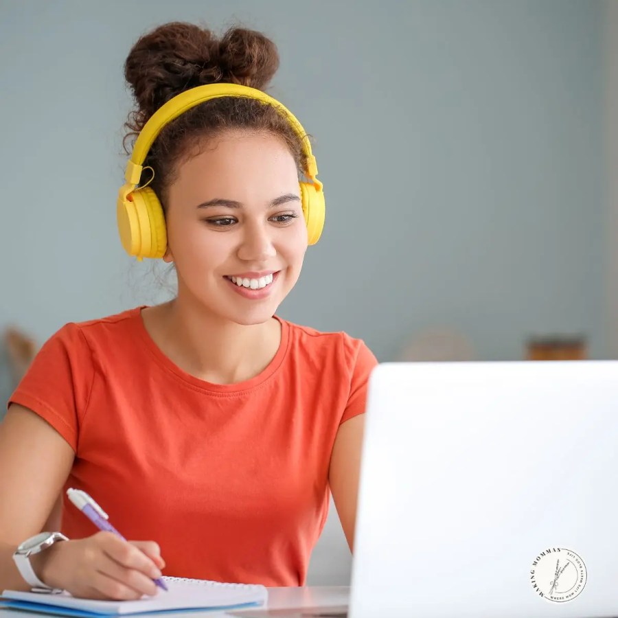 Woman wearing bright yellow headphones and an orange shirt smiles while taking notes during an online course on her laptop, representing a work-from-home mom learning new skills.