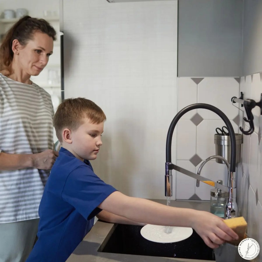 A stay-at-home mom watching her child wash dishes at the kitchen sink, showing kids helping with chores and shared responsibility at home.