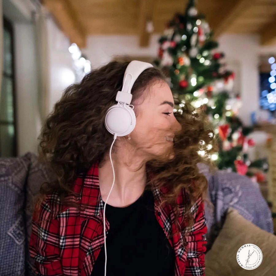 Woman wearing white headphones and smiling while listening to music in a cozy living room decorated for Christmas, symbolizing relaxation and joy for work-from-home moms during the holidays.