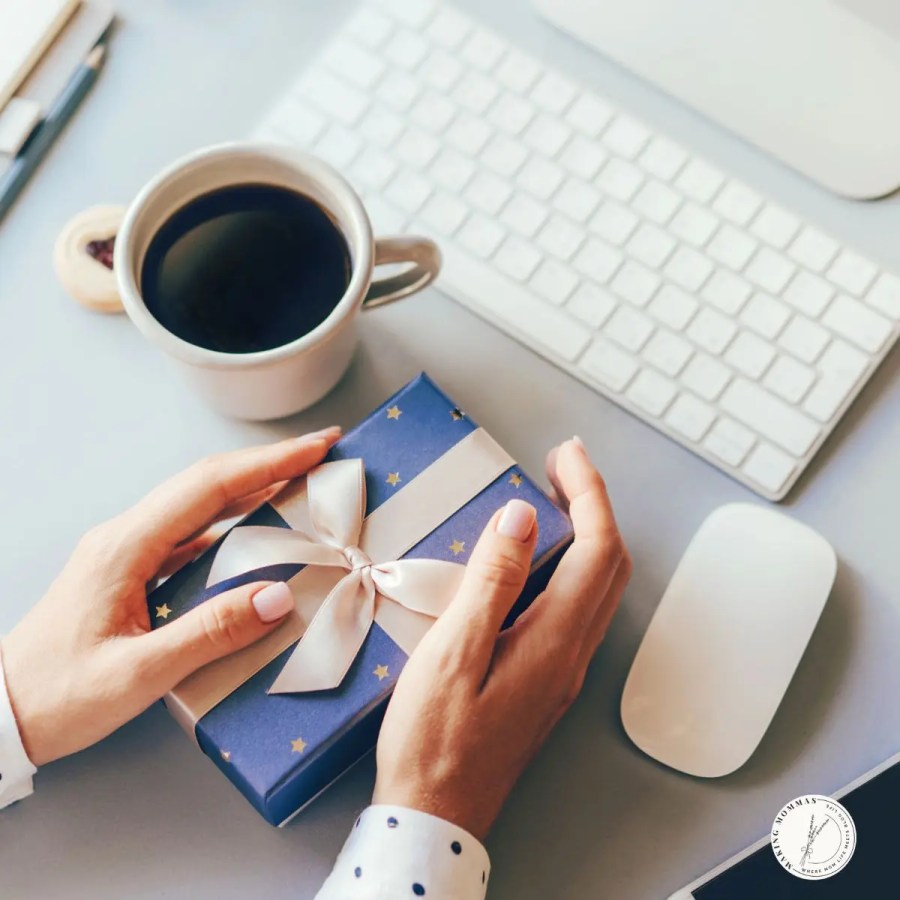 A woman's hands holding a small gift as if she is ready to open it, with a computer keyboard, mouse, and cup of coffee right by.