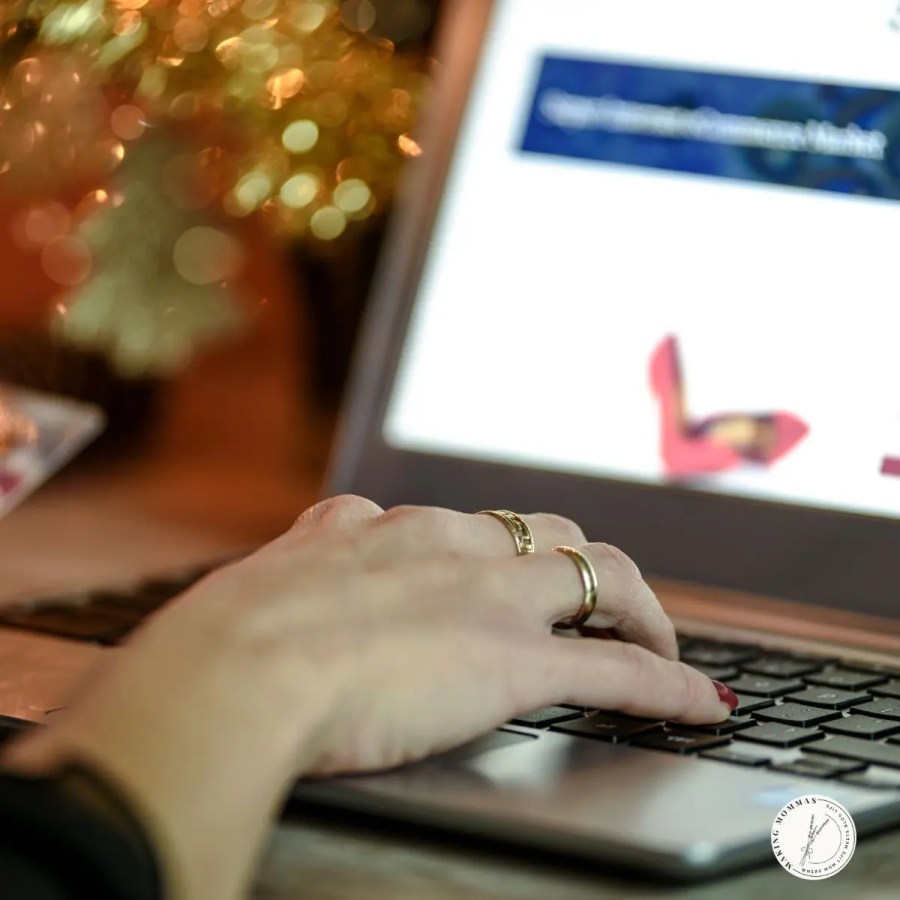 Close-up of a woman’s hand typing on a laptop keyboard with soft golden holiday lights blurred in the background, representing a cozy Christmas workspace for work-from-home moms.